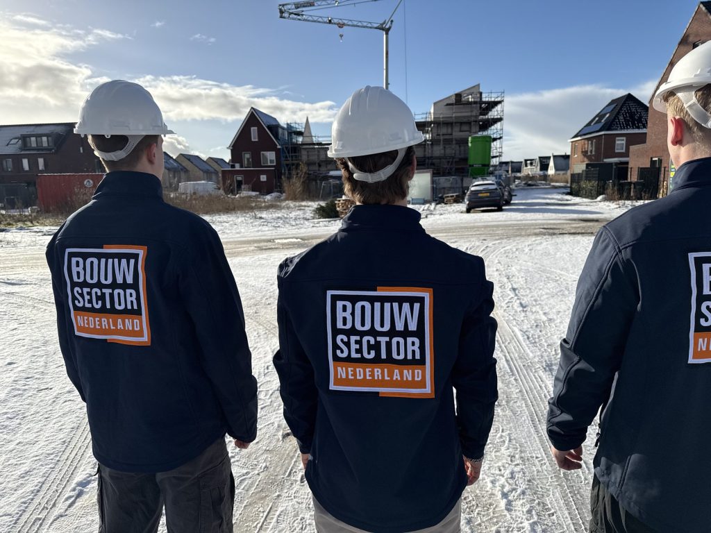 Drie vakmensen van Bouwsector Nederland met witte helm en logo op de rug, op een besneeuwde straat met nieuwbouwwoningen en een bouwkraan